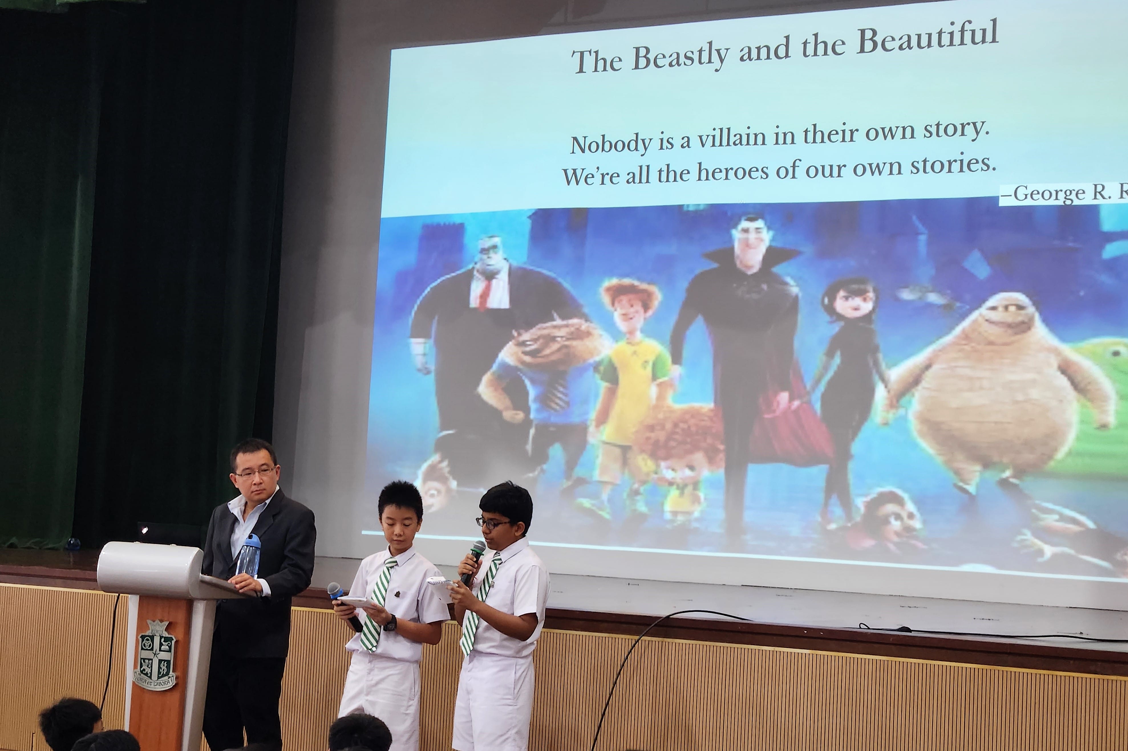 Two students holding microphones on stage beside a man at a podium, presenting a slide titled 'The Beastly and the Beautiful.'
